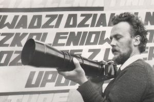 Zdjęcie czarno-białe przedstawiające mężczyznę w kręconych włosach, z brodą w ręku trzymającego aparat fotograficzny z długim teleobiektywem