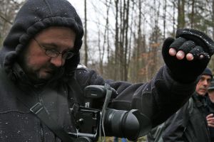 Zdjęcie przedstawiające mężczyznę w ciemnej kurtce, okularach i z kapturem na głowie, patrzącego na aparat fotograficzny stojący na statywie